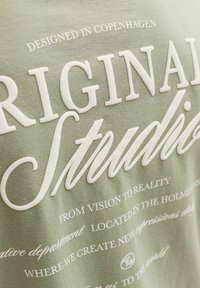 Tela verde con texto blanco en relieve que dice "Original Studios" y frases impresas más pequeñas sobre diseño y ubicación en Copenhague.