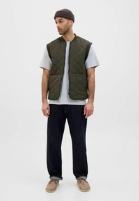 Oliven grøn, quilted vest med stående krave, båret over en grå t-shirt, parret med mørke jeans og beige fodtøj.