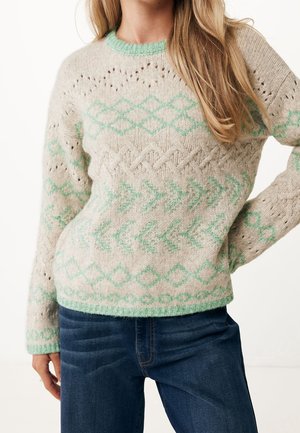 Beżowy dziany sweter z jasnooliwkowymi geometrycznymi wzorami i wzorami strzałek, w połączeniu z ciemnoniebieskimi jeansami, noszony przez osobę z długimi blond włosami.