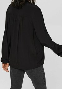Blouse noire à manches longues avec une texture douce, une couture arrière froncée et une coupe décontractée, associée à un jean gris foncé.