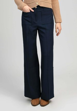 Femme portant un pantalon large bleu marine avec des poches appliquées, un cardigan en maille beige et des bottes brodées marron.