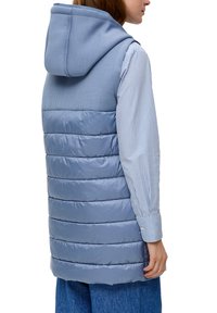 Helles blaues, gepolstertes Weste mit Kapuze, die horizontale gesteppten Abschnitte aufweist. Auf der Rückseite ist der Begriff "OLIVER" geprägt. Getragen über einem gestreiften Hemd.