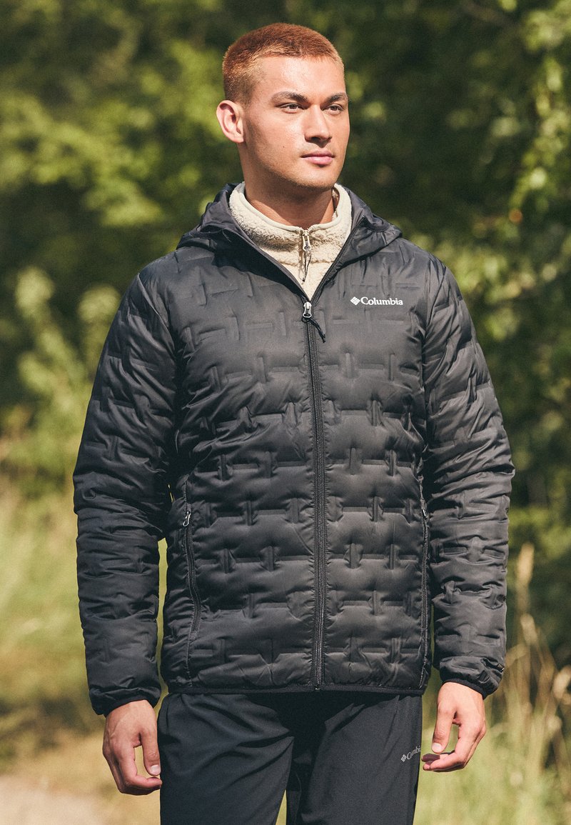 Veste matelassée noire avec fermeture éclair, dotée d'une capuche et d'un motif texturé. Logo sur la poitrine gauche, portée sur un pull en polaire de couleur claire.
