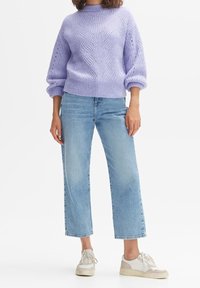 Kvinna iklädd en lavendelfärgad kabelstickad tröja, ljusblå raka jeans och vita sneakers som står mot en vit bakgrund.