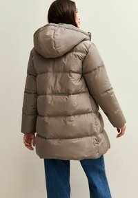 Vrouw in een beige gewatteerde pufferjack met capuchon, staand met de rug naar de camera, gecombineerd met blauwe spijkerbroeken tegen een effen lichte achtergrond.