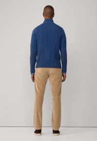 Blauwe kabelgebreide trui met een hoge kraag, gedragen met beige corduroy broek en bruine schoenen, gezien van achteren.
