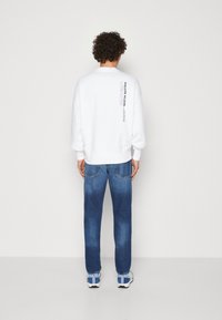 Sudadera blanca con cuello redondo y logo en la parte trasera. Combinada con jeans de mezclilla azul, que presentan un corte recto y un acabado desgastado.