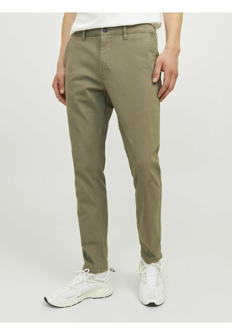 Homme portant un pantalon slim vert olive et des baskets blanches, debout devant un fond clair uni.