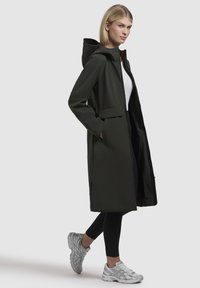 Cappotto lungo impermeabile di colore verde scuro, con cappuccio, tasche laterali e vestibilità comoda, abbinato a leggings neri e sneakers grigio chiaro.