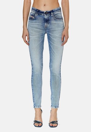 Femme portant un jean skinny taille haute bleu clair et des talons ouverts en denim, debout contre un fond blanc, les mains détendues le long du corps.