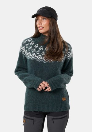 Donna che indossa un maglione di lana verde scuro con motivo nordico bianco, pantaloni grigio scuro con tasche con cerniera e cappellino da baseball nero, mani intrecciate.
