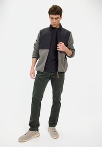 Grau und schwarze Zip-Jacke mit Stehkragen, kombiniert mit dunkelgrünen Hosen und hellbraunen Sneakers. Glatte Stofftextur und lässiges Design.
