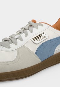 Κοντινή λήψη λευκού αθλητικού παπουτσιού Puma Palermo με μπλε πλευρική ρίγα, γκρι κορδόνια, καφέ σόλα και ορατό λογότυπο Puma στο πλάι.