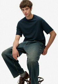 Jonge man met bruin haar zit op een kruk en draagt een donkerblauw t-shirt, losse vervaagde spijkerbroeken, grijze sokken en bruine leren schoenen.