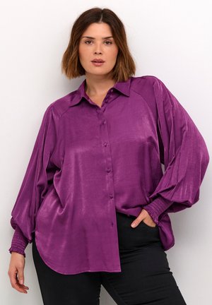 Camisa de satén morado de botones con corte holgado, mangas largas abullonadas y puños fruncidos. Presenta un cuello con solapa y un dobladillo redondeado.