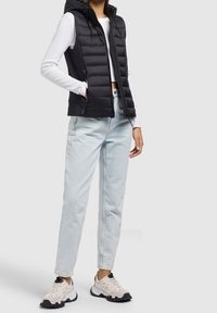 Schwarze Steppweste mit Kapuze und Reißverschlusstaschen, getragen über einem weißen Langarmshirt. Hellblaue Straight-Leg-Jeans und Sneakers mit pinken Akzenten.