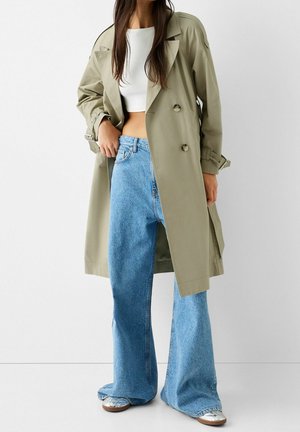 Kobieta w beżowym trenczu na białym crop topie, szerokie niebieskie jeansy i srebrne sneakersy, stojąca na jednolitym tle.