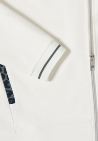 Manche de veste blanche zippée avec un poignet côtelé présentant une bande sombre unique et un bord de poche à motif camouflage visible.