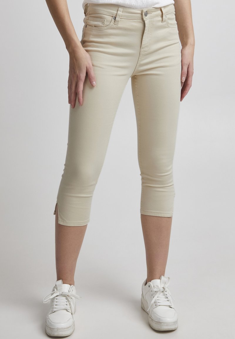 Jasnobeżowe spodnie capri wykonane z elastycznego materiału, z średnim stanem, przednimi kieszeniami i bocznymi rozcięciami u dołu. Połączone z białymi sneakersami.