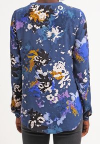 Långärmad blus i blått tyg med ett blommönster i vitt, svart, lila och guld. Rak skärning med rundad söm bak.