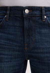 Dunkelblaue Jeans mit tailliertem Schnitt, silberner Knopfverschluss und kontrastierenden orangen Nähten. Verfügt über fünf Taschen und eine glatte Textur.