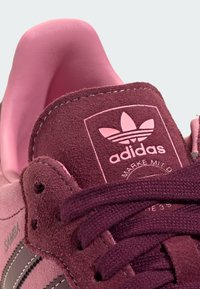 Nahaufnahme der Zunge des Adidas Samba-Sneakers aus pinkem und kastanienbraunem Veloursleder mit Logo, Nähten und gebundenen Schnürsenkeln.