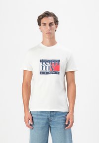 Mand med kort krøllet hår iført en hvid Tommy Jeans t-shirt og lys blå jeans, stående mod en ensfarvet hvid baggrund.