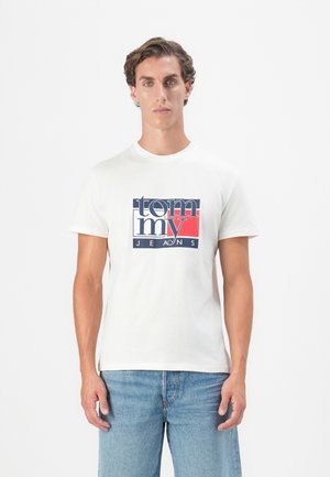 Homme aux cheveux courts et bouclés portant un t-shirt blanc Tommy Jeans et un jean bleu clair, se tenant devant un fond blanc uni.