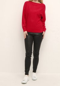 Pull en maille rouge avec un large décolleté, pantalon en cuir noir ajusté, et baskets blanches, avec une coupe décontractée et des poignets côtelés.