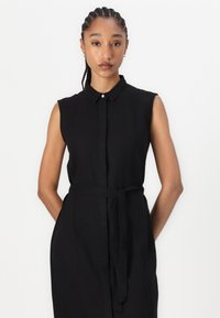 Svart, ärmlös klänning med krage som knäpps med knappar, cinchad midja med ett matchande bälte och sidofickor. Slät tyg med minimalistisk design.