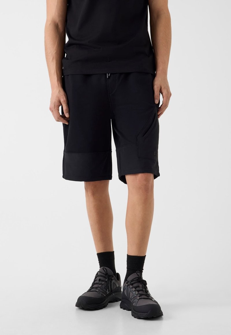 Mann trägt schwarze knielange Cargo-Shorts, schwarze knielange Socken und schwarze Sportschuhe, steht vor einem schlichten hellen Hintergrund.