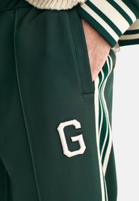 Zielone joggery z gładkiego materiału, ozdobione białym haftowanym litery "G" oraz paskami w odcieniu kremowym i zielonym wzdłuż boków.