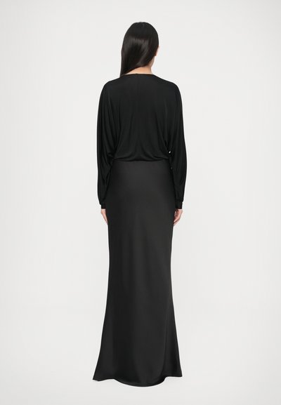 Sieviete ar gariem tumšiem matiem stāv sejā prom, valkājot melnu garām piedurknēm blūzi un melnu grīdas garuma svārku vienkāršā fonā.