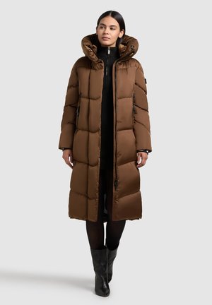 Hnědý puffák s kapucí, prošívaný design, zapínání na zip a šňůrky. Modelka ho nosí přes černý rolák a tmavé legíny.