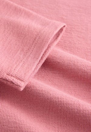 Artimas rožinės mezginio audinio, sulankstyto, vaizdas, kuriame matyti tekstūruotas pynimo raštas ir siūlių detalė krašte.