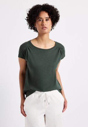 Donkergroen t-shirt met korte mouwen en een ronde halslijn, voorzien van subtiele plooien op de schouders. Gekozen in combinatie met witte broek met trekkoord.