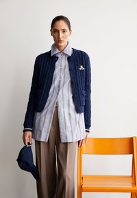 Dunkelblauer strukturierter Cardigan mit Knöpfen, kombiniert mit einem hellblauen gestreiften Hemd und beigen weiten Hosen. Dunkelblake Mütze in der Hand.