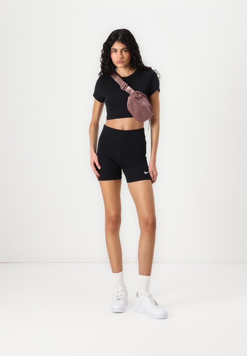 T-shirt noir raccourci et shorts taille haute, associés à un sac bandoulière rose clair et des baskets blanches avec des lacets texturés.
