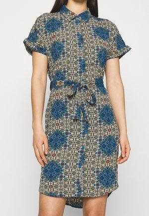 Kvinde iført kortærmet, knap-op kjole med blåt og brunt geometrisk blomsterprint og bundet stofbælte i taljen.