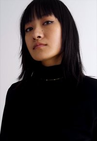 Schwarzes Rollkragenoberteil, das eine zarte goldene Halskette mit kleinen Perlen zeigt, modelliert von einer Person mit glattem schwarzen Haar und minimalem Makeup.