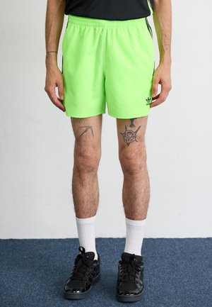 Neon green athletic shorts with elastic waistband, side black stripes, and adidas logo. Worn with black sneakers and white socks.