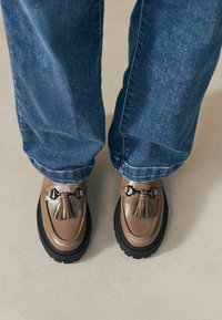 Bruna läderskor med tofsar, svarta kraftiga sulor, i kombination med blå denimjeans. Tydlig textur och sömmar på skorna.