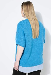 Maglione azzurro di grandi dimensioni a maniche corte, realizzato in un tessuto testurizzato. Indossato sopra una camicia a righe bianca e blu.