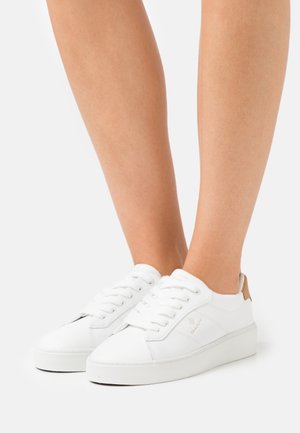 Jambes portant des baskets blanches basses avec des lacets et un onglet de talon marron, debout sur un fond clair uni.
