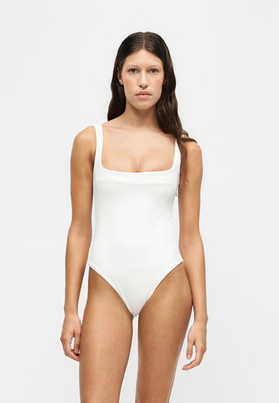 Γυναίκα με μακριά σκούρα μαλλιά, φοράει λευκό αμάνικο ολόσωμο μαγιό και στέκεται μπροστά σε απλό ανοιχτό φόντο.