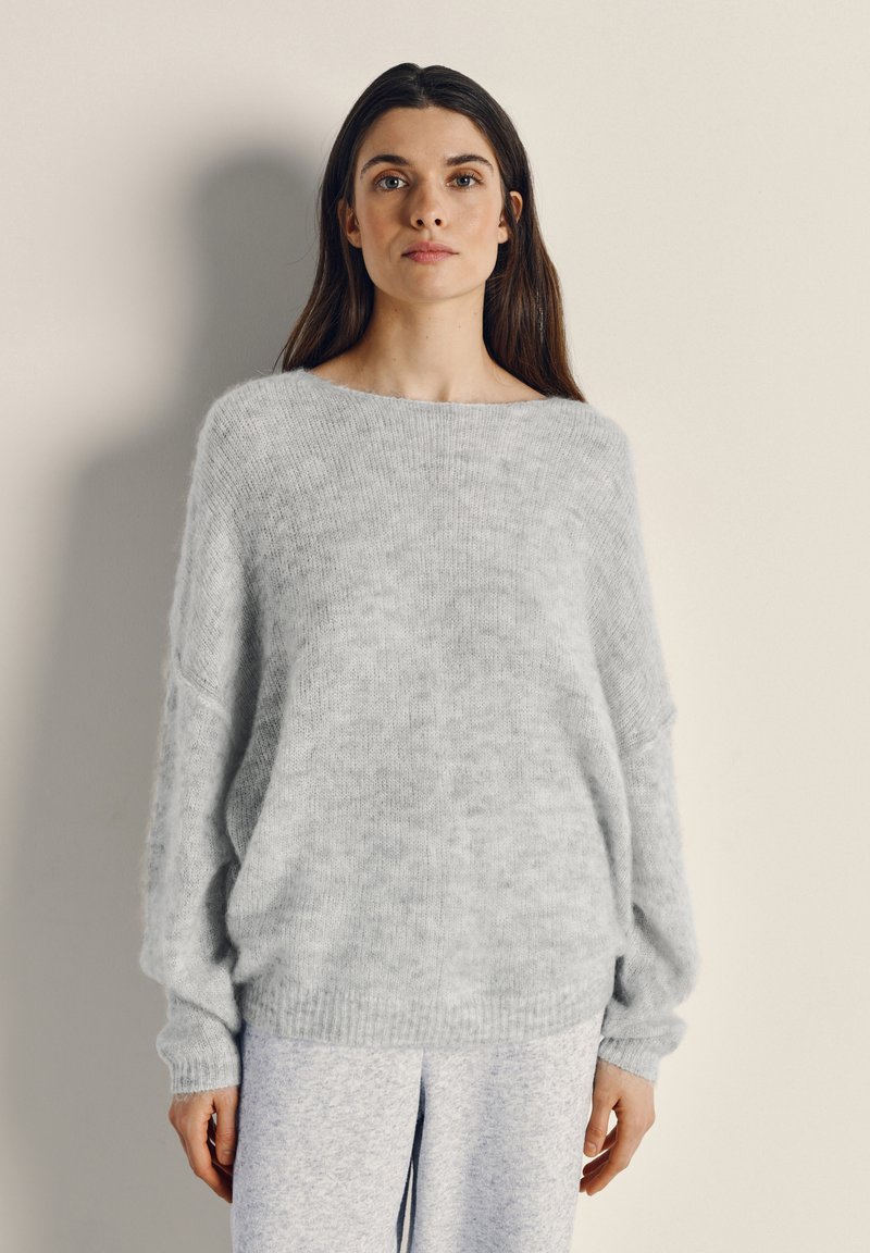 Žena oblečená v ľahkom sivom chlpatom svetri a zodpovedajúcich nohaviciach, stojaca pred jednoduchým béžovým pozadím, čelom vpred s neutrálne výrazom.