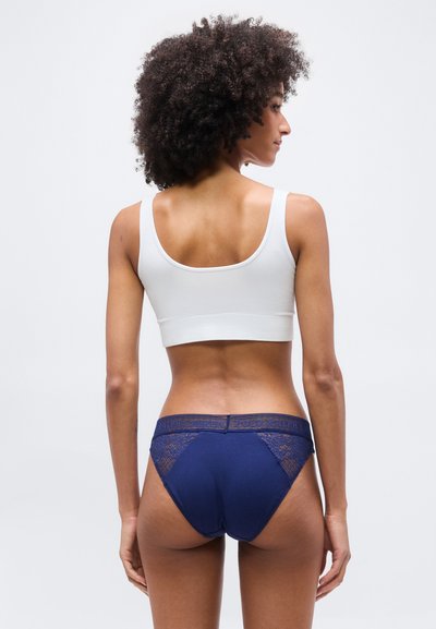 Γυναίκα με σγουρά μαλλιά, φορώντας λευκό κοντό τοπ και εσώρουχα μπλε σκούρα με δαντέλα, στέκεται με την πλάτη στην κάμερα.