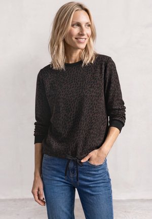 Smilende kvinde med blondt hår iført en mørk mønstret langærmet top og blå jeans, poserer med den ene hånd i lommen.