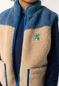 Kind mit einer blauen und beigen Fleece-Weste mit blauen Akzenten und einem grünen und beigen Logo auf der Brust, über einem navyblauen Langarmshirt.