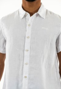 Camicia corta bianca in lino con colletto button-down, cinque bottoni e un logo ricamato sul petto, caratterizzata da un tessuto texture.